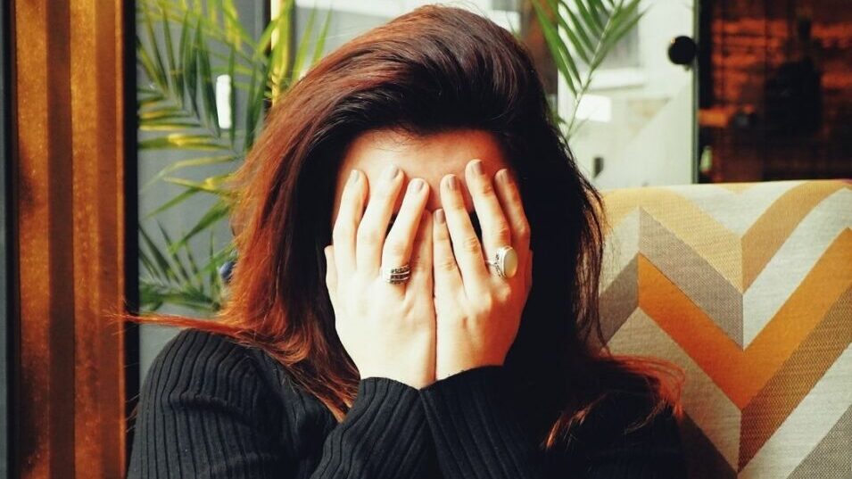 Person covering face in cozy café chair with patterned upholstery and plants in the background.